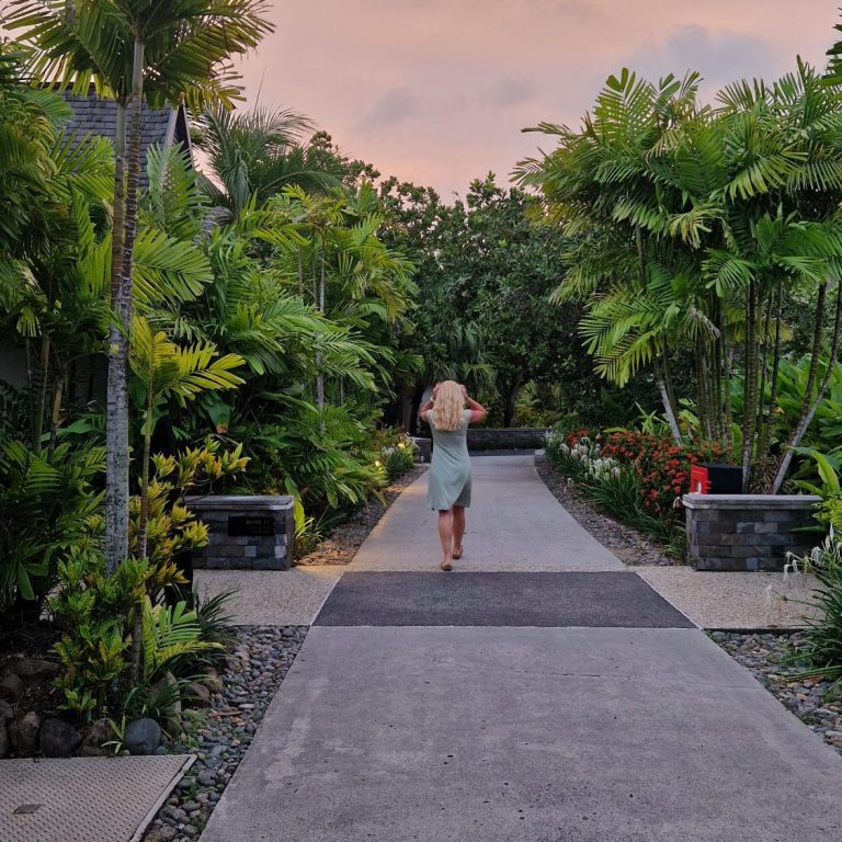 Eine Frau in einem grünen Kleid geht auf einem Weg, umgeben von tropischen Pflanzen.