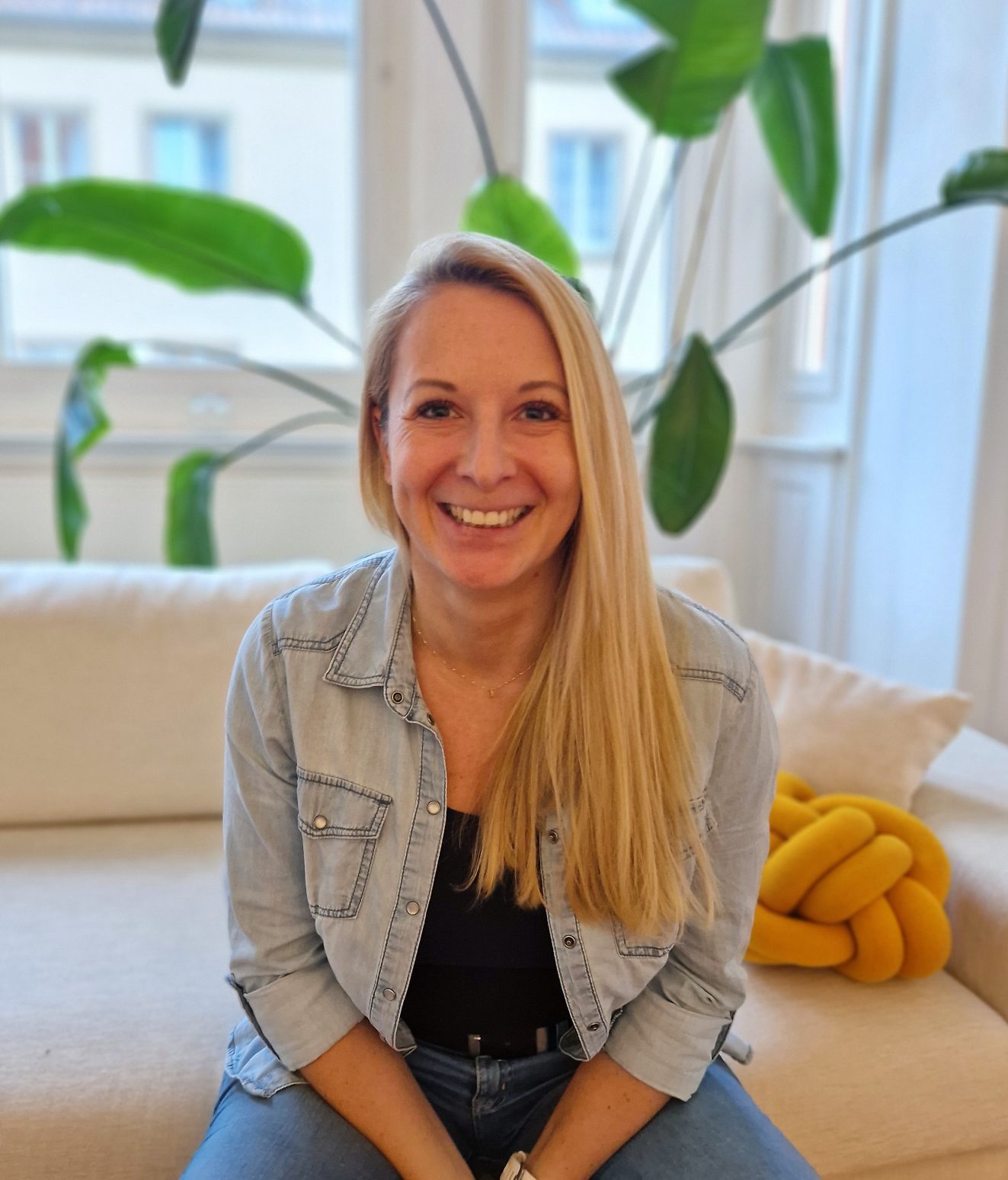 Lächelnde Frau mit langen blonden Haaren sitzt auf einer Couch, umgeben von Pflanzen.